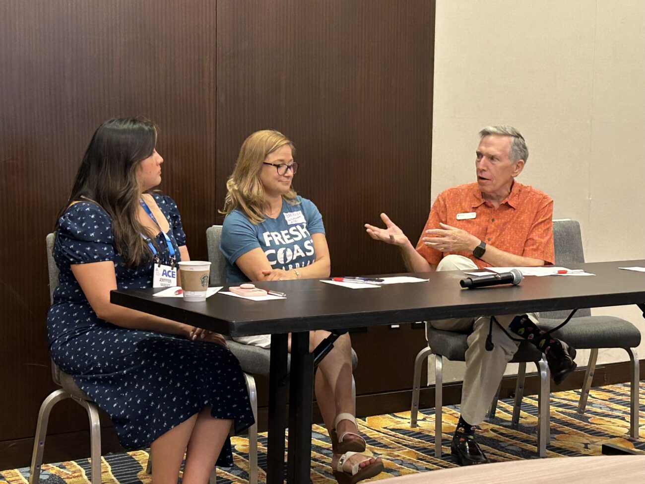 Three people sit around a conference table in a panel discussion, with papers, a microphone, and cups in front of them.