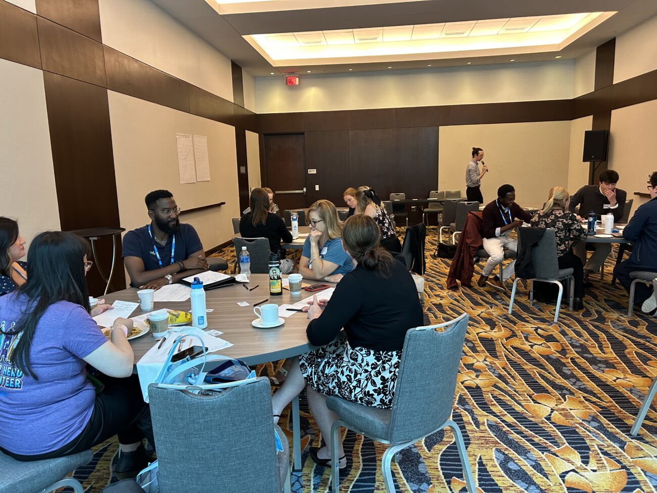 A conference room with people seated at round tables, eating and chatting, while a man with a microphone stands at the front.