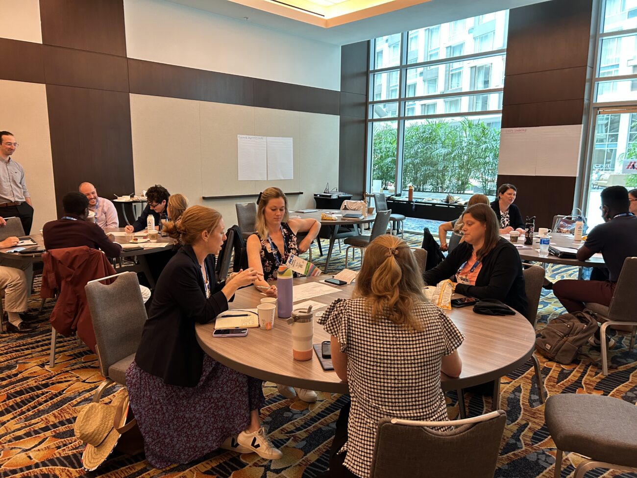 Professionals seated around round tables in a bright conference room, taking notes and chatting during a meeting.