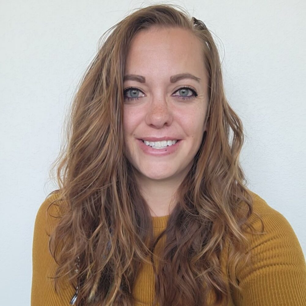 Smiling woman with long wavy light brown hair wearing a mustard sweater, against a light background.