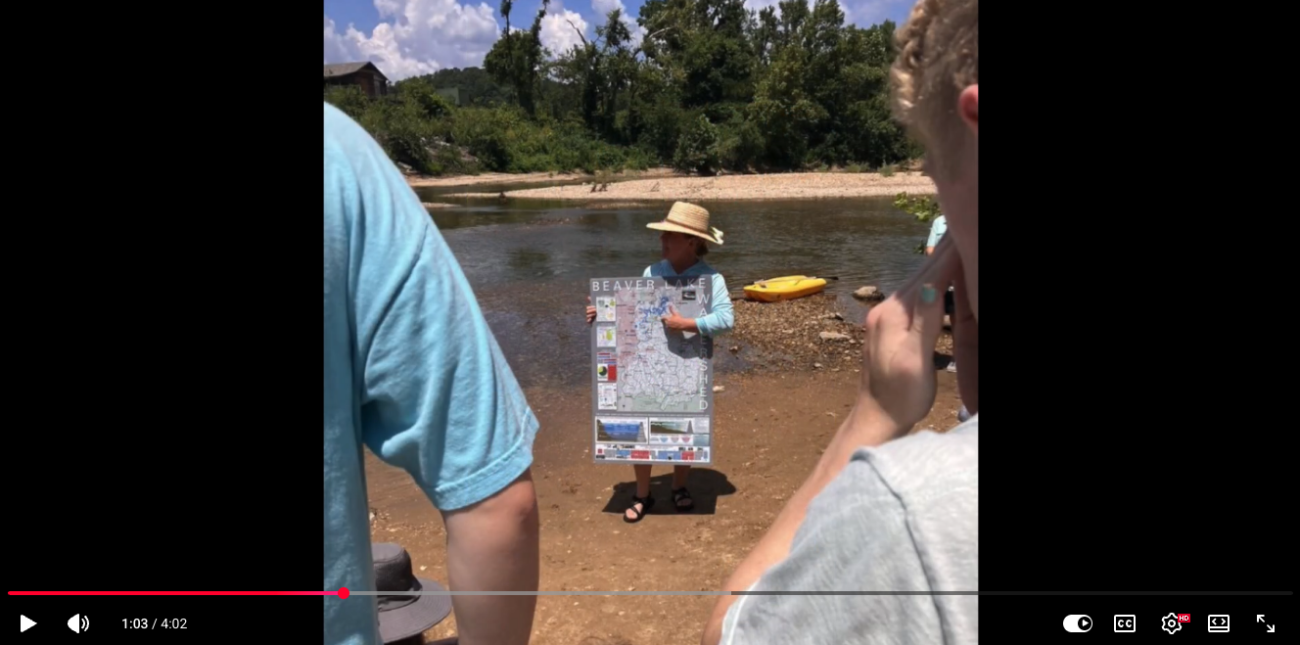 Person wearing a straw hat holds a large map poster on a riverbank while a small group watches nearby.
