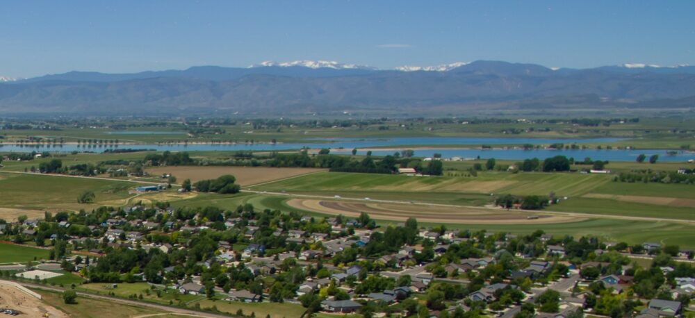 Aerial view of a town with green lawns and houses, surrounded by farmland, a blue river, and distant snow-capped mountains.