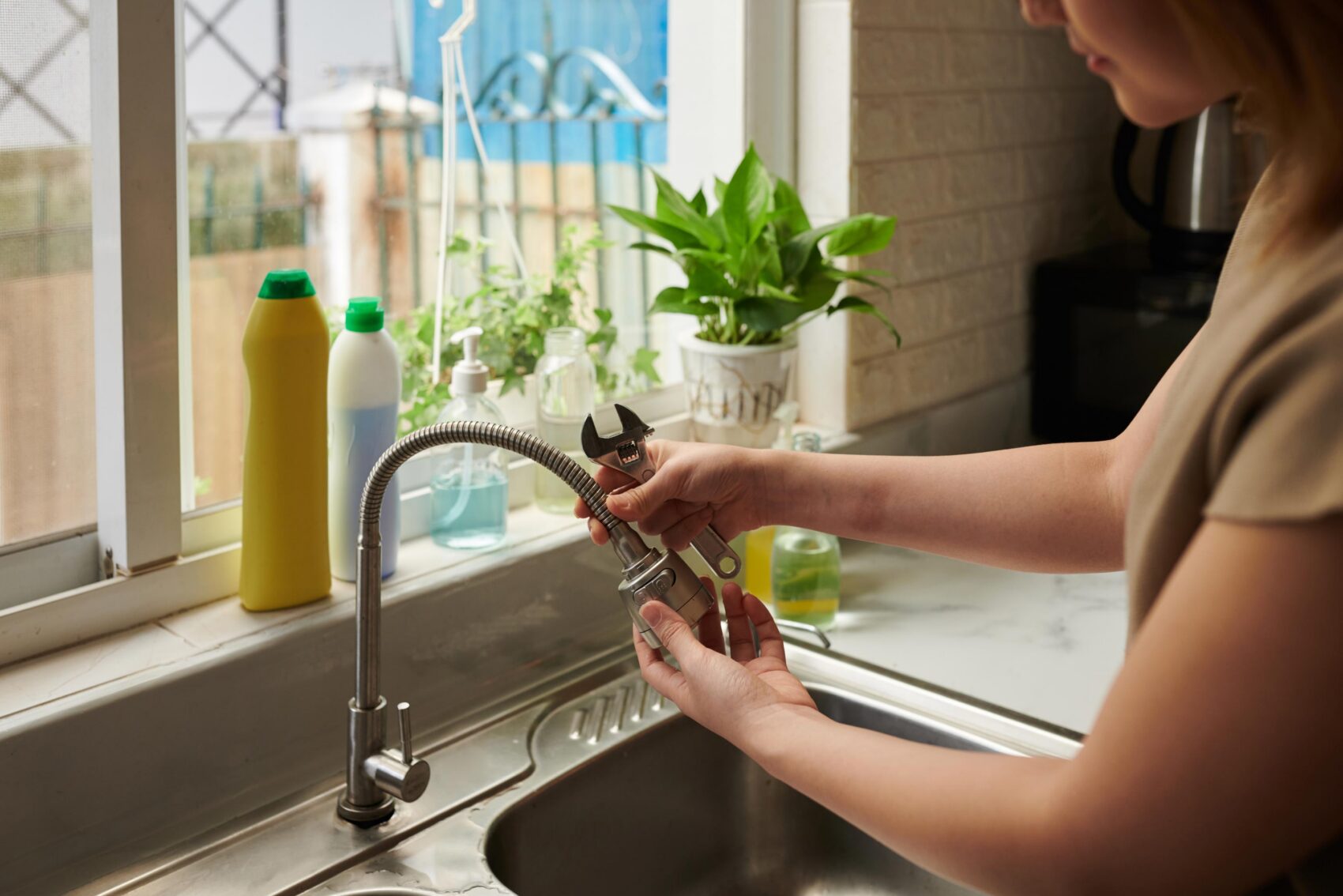 Person using a wrench to repair a kitchen sink faucet, with dish soap bottles and a potted plant on the windowsill behind them.