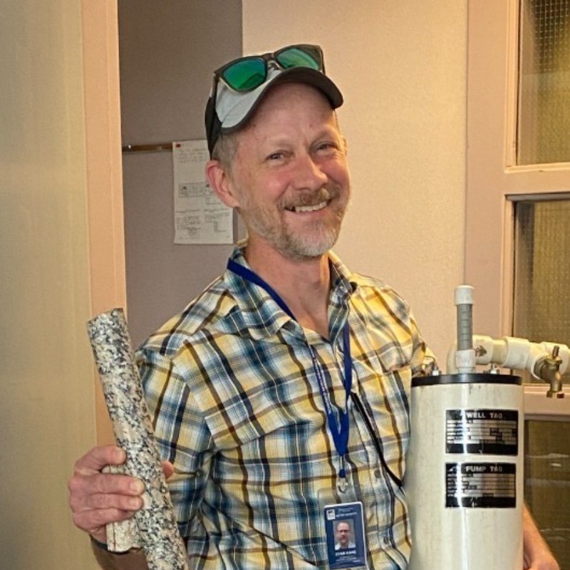 Smiling man in a plaid shirt and cap, wearing a badge, holds a rolled poster in one hand and a water-filter unit in the other in a workshop setting.