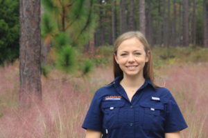 Smiling woman in a navy uniform standing in a pine forest with pink grass in the foreground.