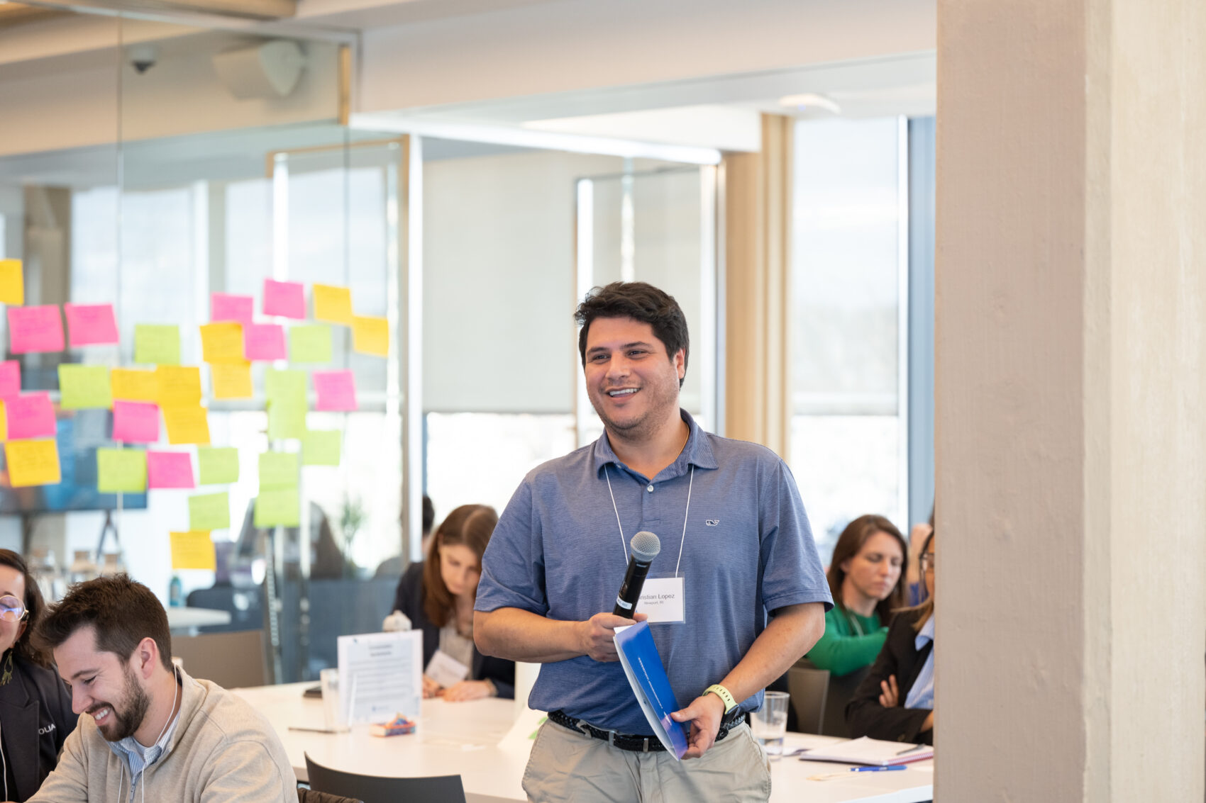 Man standing and speaking into a handheld microphone at a busy desk with colorful sticky notes on glass walls behind him.