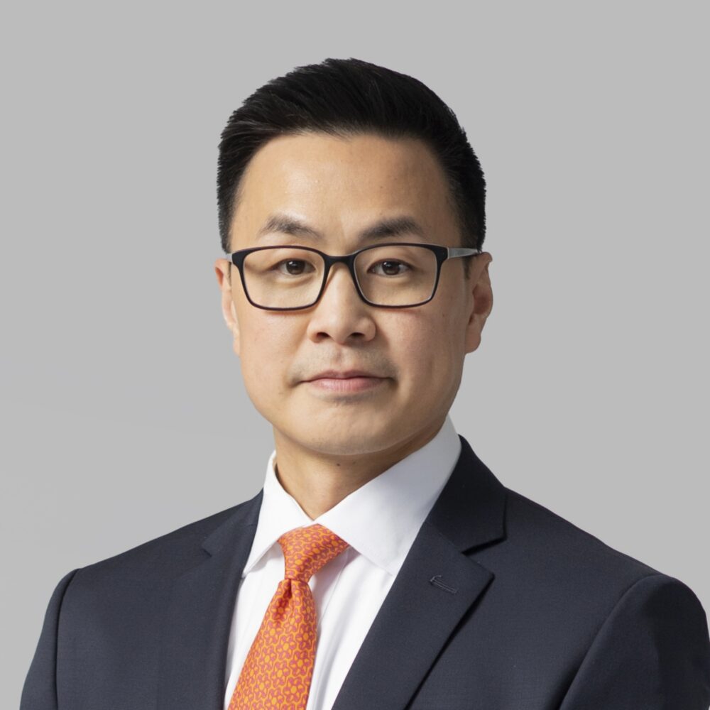 Portrait of a man wearing glasses, a navy suit, white shirt, and an orange patterned tie against a gray background.