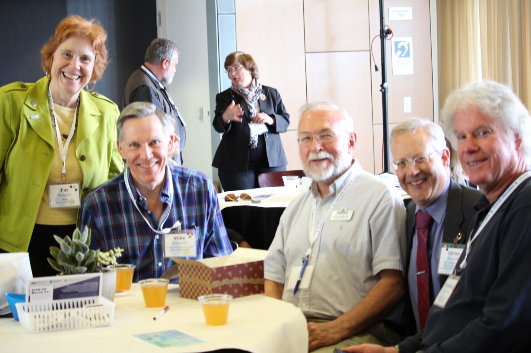 Smiling group of professionals at a conference table with name badges and snacks in a casual networking moment.