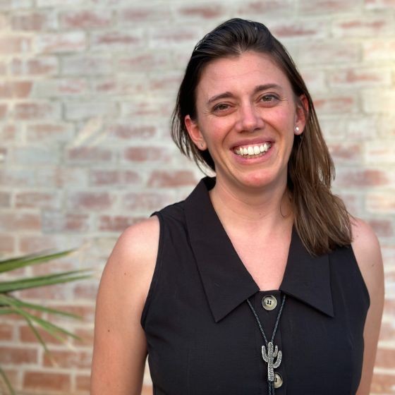 Smiling woman with brown hair in a black sleeveless blouse, wearing a cactus pendant, standing in front of a brick wall.