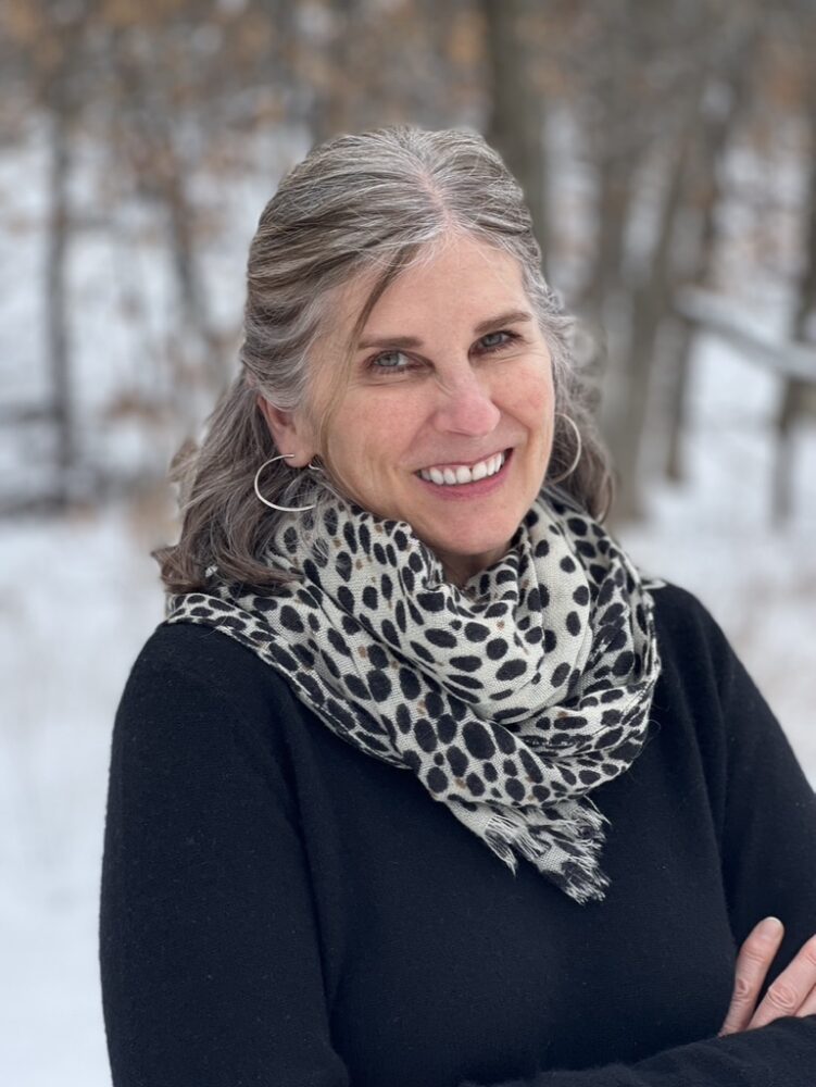 Smiling woman with gray hair wearing a leopard-print scarf and black sweater outdoors in snowy woods.