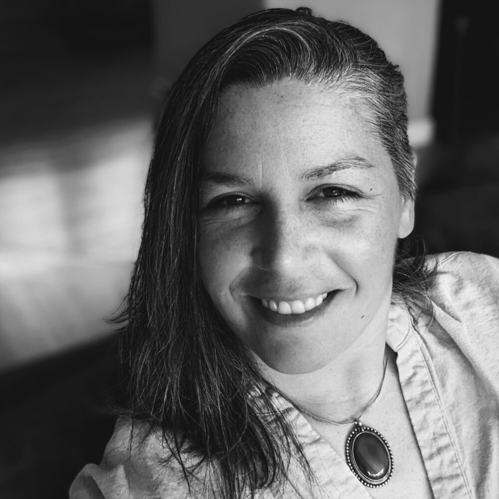 Close-up black-and-white portrait of a smiling woman, showing eyes, nose, and a large oval pendant necklace partially visible on her chest.