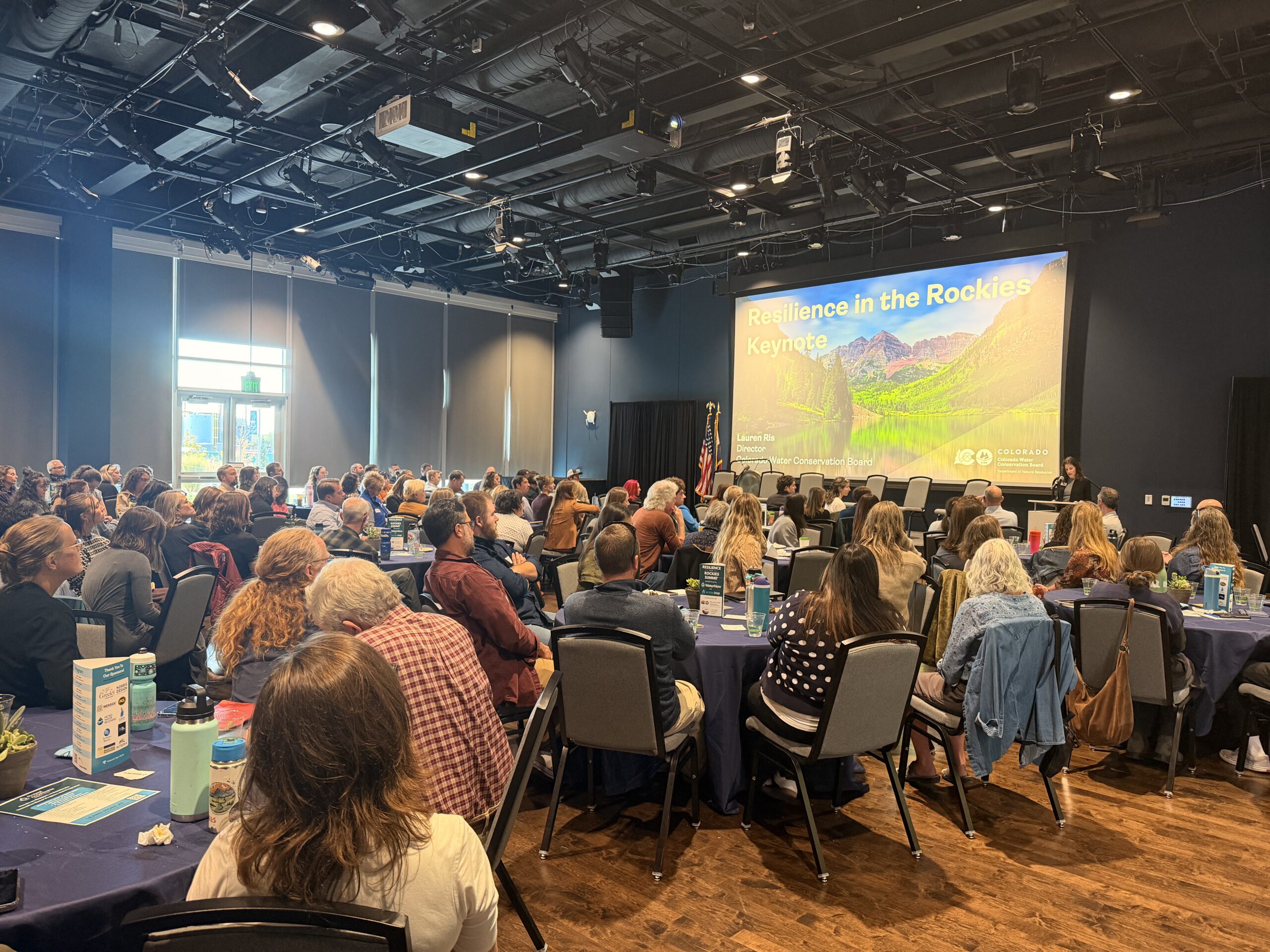 Audience seated at round tables facing a stage with a large screen reading 'Resilience in the Rockies Keynote' and a mountain landscape backdrop.