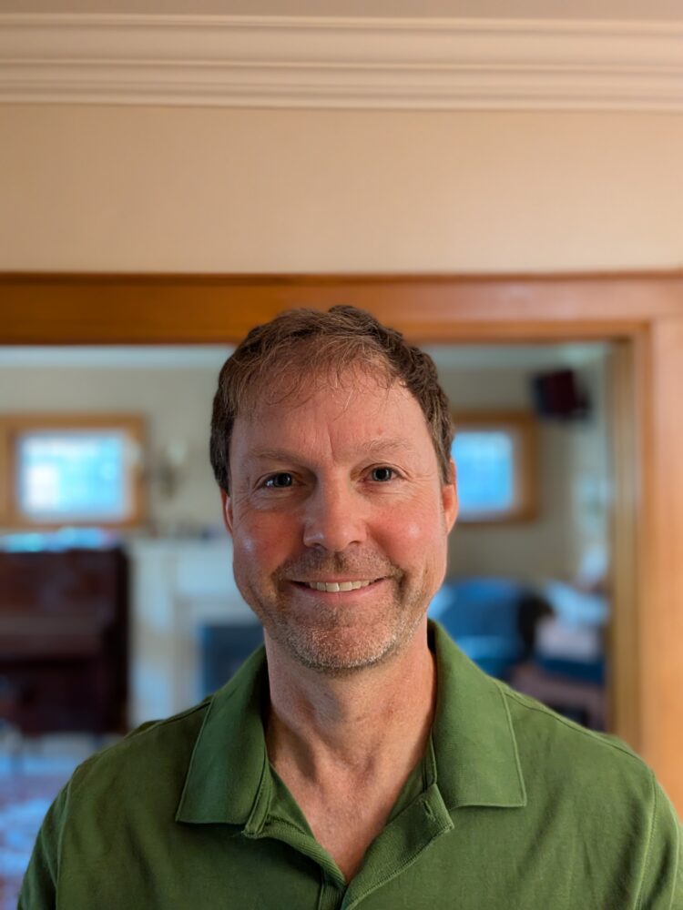 Smiling man in a green polo shirt indoors, wood trim and blurred windows in the background.