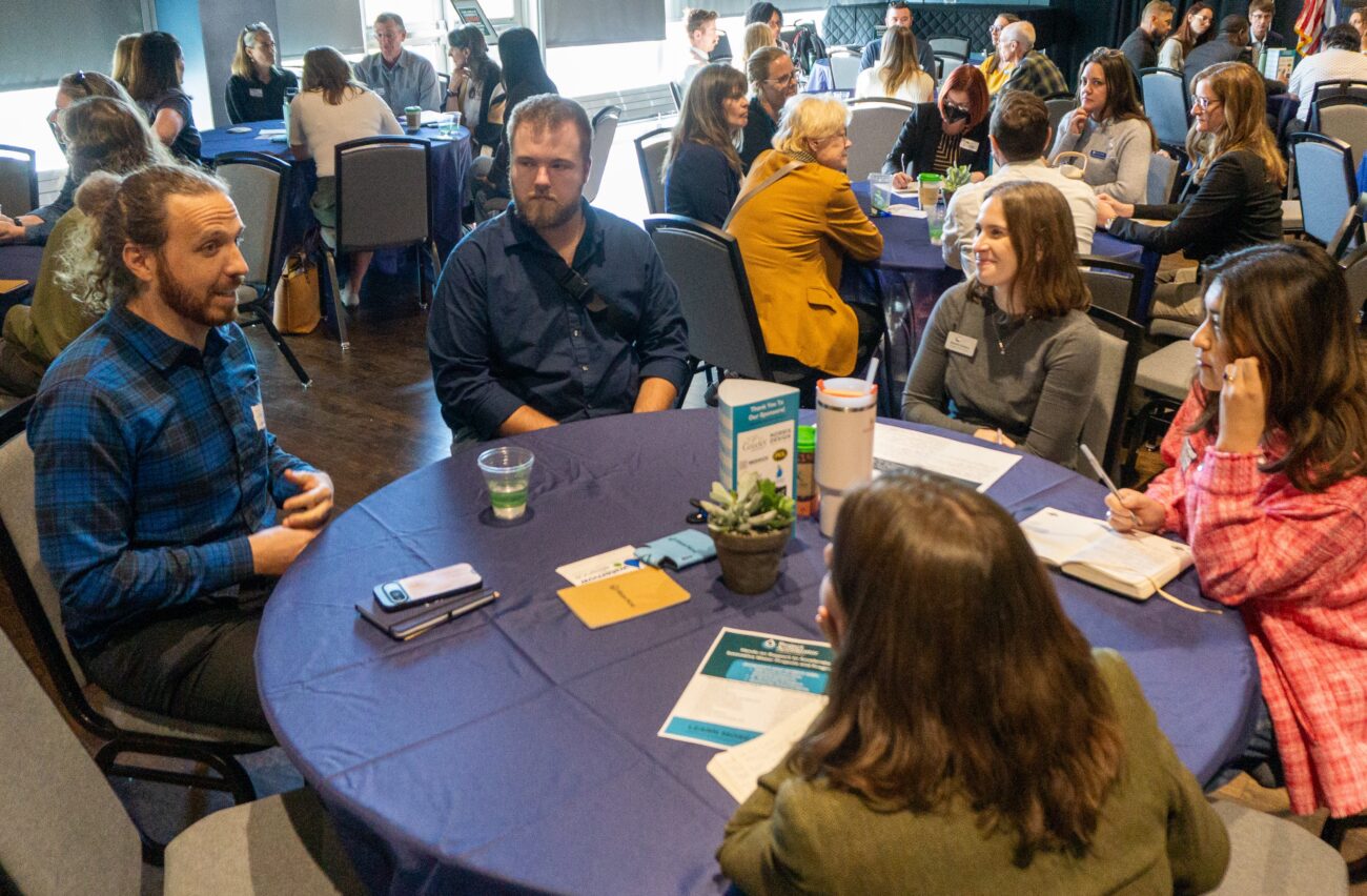 People seated around round conference tables talking and taking notes; blue tablecloths, name badges, cups, and a small plant at the center of a table.