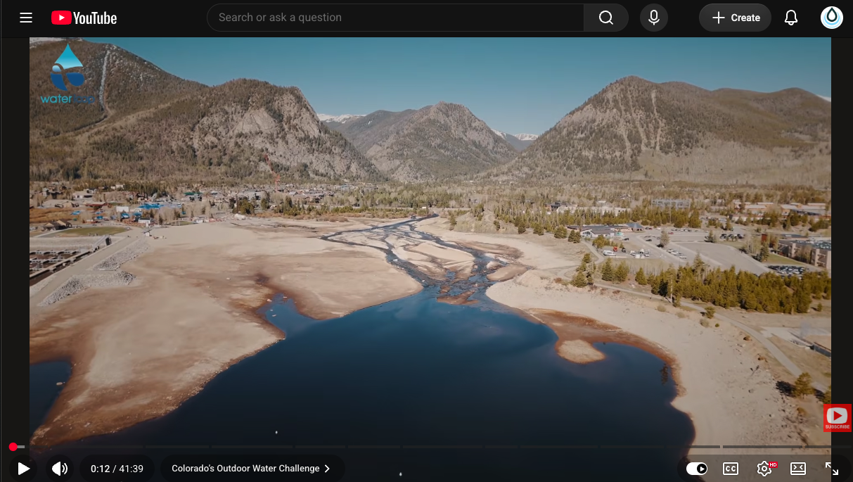 Aerial view of a winding river through a dry brown riverbed toward a blue lake, with mountains and a small town in the distance and a water-drop logo in the top-left corner.