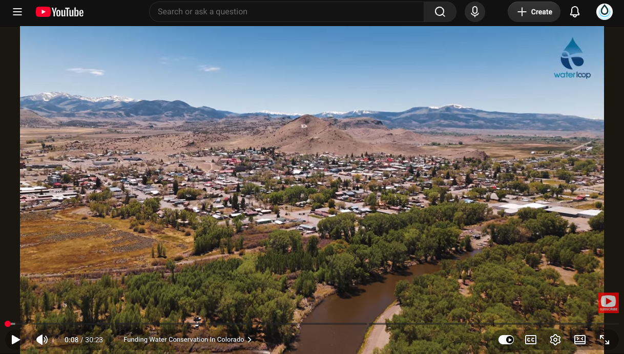 Aerial view of a small town along a winding river, with green trees and brown hills, and distant snow-capped mountains under a clear blue sky; WaterLoop logo in the top-right.